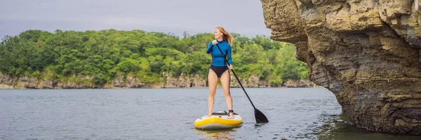 BANNER, LANG FORMAT Jonge vrouwen die plezier hebben staan te peddelen in de zee. Een SUP. Rood haar meisje training op peddel boord in de buurt van de rotsen — Stockfoto