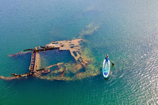 Frau auf Paddelbrett, neben verlassenem Schiffswrack, das aus dem Meer ragt — Stockfoto