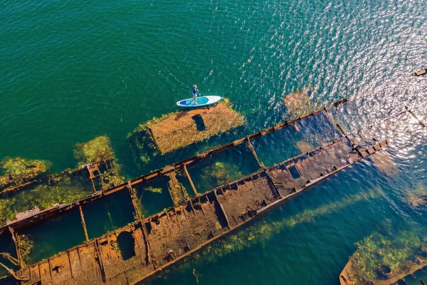 Femme sur la planche à pagaie, soup à côté de naufrage cassé abandonné sortant de la mer — Photo