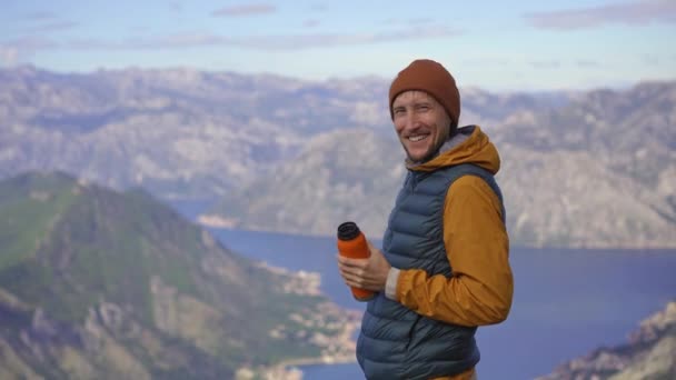 Een man reist in het najaar in Montenegro. Hij drinkt een warm drankje uit een thermoskan terwijl hij op een uitkijkpunt staat met uitzicht op de Boka Kotorska baai. — Stockvideo