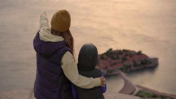 Um tiro à mão. Uma mulher e seu filho viajantes visitam o miradouro sobre a ilha Sveti Stefan. Famosa localização turística perto da cidade de Budva. Viagem de outono a Montenegro — Vídeo de Stock