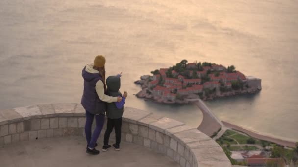 Um tiro à mão. Uma mulher e seu filho viajantes visitam o miradouro sobre a ilha Sveti Stefan. Famosa localização turística perto da cidade de Budva. Viagem de outono a Montenegro — Vídeo de Stock