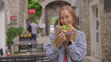 Genç bir kadın, eski bir Avrupa şehrinin caddesinde duran siyah ekmek üzerinde lezzetli bir pizza yiyor.