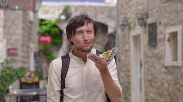 Ein junger Mann isst eine leckere Pizza auf Schwarzbrot, die auf einer Straße einer alten europäischen Stadt steht — Stockvideo