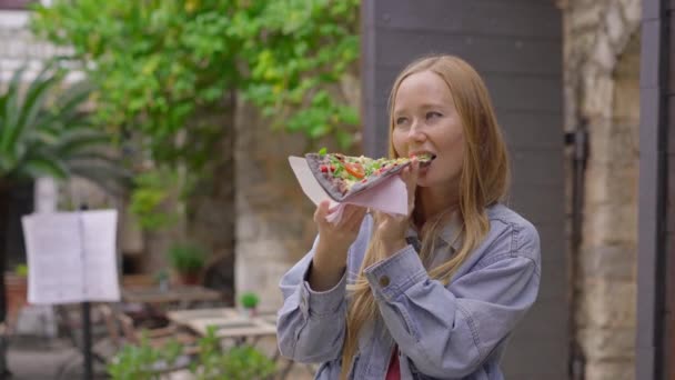 Una joven come una deliciosa pizza sobre pan negro de pie en una calle de un antiguo pueblo europeo — Vídeos de Stock