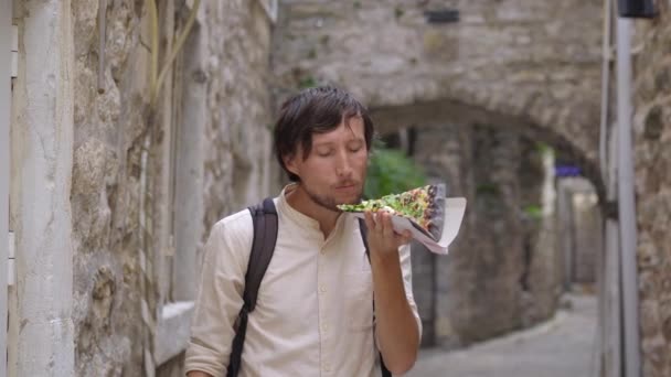 A young man eats a delicious pizza on black bread standing on a street of an old European town — Wideo stockowe