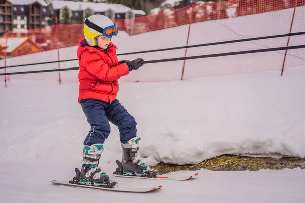 Le garçon utilise un ascenseur d'entraînement. Ski d'enfant en montagne. Enfant tout-petit actif avec casque de sécurité, lunettes et bâtons. Course de ski pour jeunes enfants. Sport d'hiver pour la famille. Cours de ski pour enfants à l'école alpine — Photo