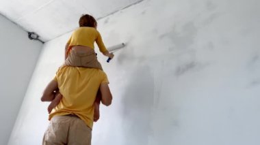 Sarı tişört giyen bir baba ve oğul, duvarları tekerlekli boyayla boyarlar. Dairelerini yeniliyorlar. DIY ev yenileme konsepti. Aile zamanı. Yavaş çekim.
