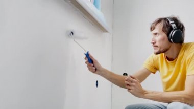 Sarı tişört giyen bir adam paten boyacısı kullanarak duvarı boyar. O, kulaklıklarıyla müzik dinleyerek dairesini yeniliyor. DIY ev yenileme konsepti. Yavaş çekim.