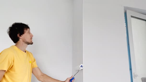 Mężczyzna w żółtej koszulce maluje ścianę za pomocą malarza wałków. Remontuje swoje mieszkanie. Koncepcja renowacji domu DIY. Strzał zwolniony — Wideo stockowe