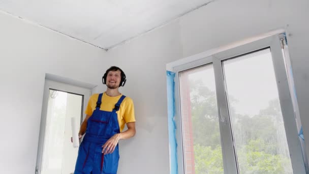 Een professionele bouwvakker schildert de muur met een rolschilder. Hij luistert naar muziek in z 'n koptelefoon. — Stockvideo