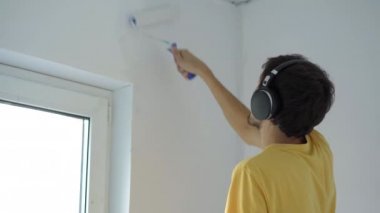 Sarı tişört giyen bir adam paten boyacısı kullanarak duvarı boyar. O, kulaklıklarıyla müzik dinleyerek dairesini yeniliyor. DIY ev yenileme konsepti
