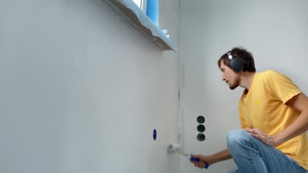 Ένας άντρας που φοράει ένα κίτρινο μπλουζάκι ζωγραφίζει τον τοίχο χρησιμοποιώντας έναν πίνακα ζωγραφικής. Ανακαινίζει το διαμέρισμά του ακούγοντας μουσική στα ακουστικά του. DIY έννοια ανακαίνιση του σπιτιού. Σφηνάκι αργής κίνησης — Αρχείο Βίντεο