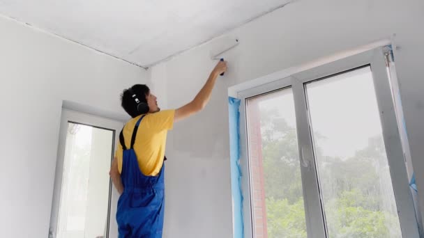 Een professionele bouwvakker schildert de muur met een rolschilder. Hij luistert naar muziek in z 'n koptelefoon. — Stockvideo