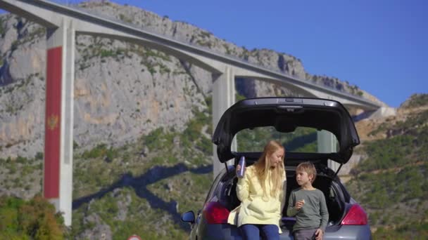 Mężczyzna i jego syn siedzą i jedzą w samochodzie z mostem Moracica w tle. Wyjeżdżają samochodem przez Czarnogórę. — Wideo stockowe