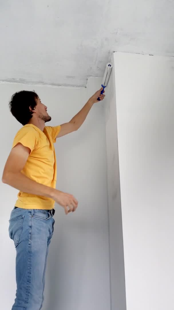 Video verticale. Un uomo che indossa una t-shirt gialla dipinge il muro usando un pittore a rullo. Sta ristrutturando il suo appartamento. fai da te concetto di ristrutturazione casa. Colpo di rallentamento — Video Stock