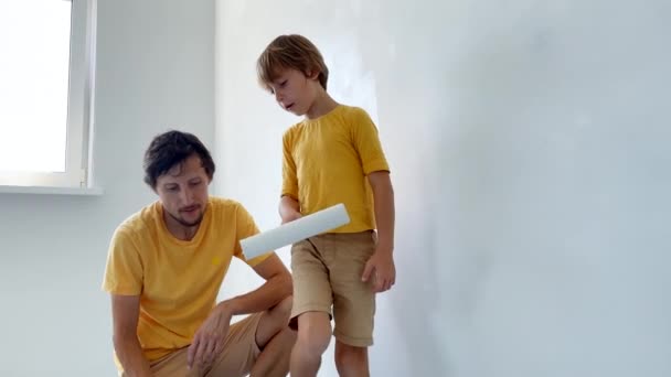 Un padre e un figlio che indossano entrambi una t-shirt gialla dipingono il muro usando un pittore a rullo. Stanno ristrutturando il loro appartamento. fai da te concetto di ristrutturazione casa. Tempo in famiglia. Colpo di rallentamento — Video Stock