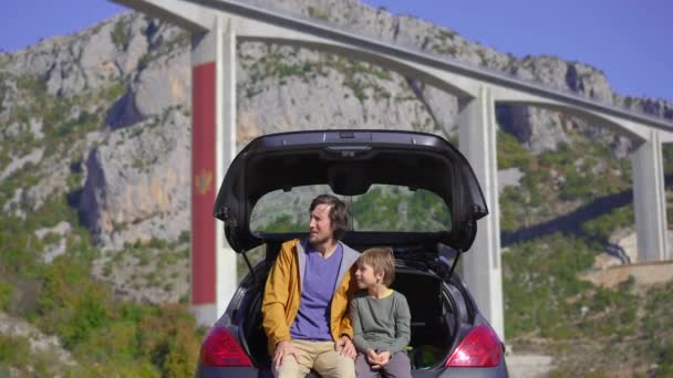 Ein Mann und sein Sohn sitzen in ihrem Auto und unterhalten sich mit der Moracica-Brücke im Hintergrund. Sie machen eine Autofahrt durch Montenegro — Stockvideo
