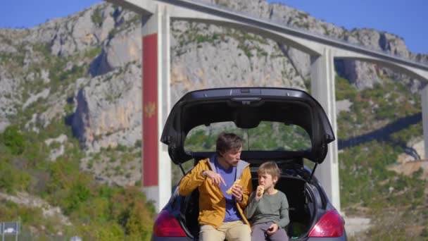 Un homme et son fils sont assis et mangent dans leur voiture avec le pont Moracica en arrière-plan. Ils font un voyage en voiture à travers le Monténégro — Video