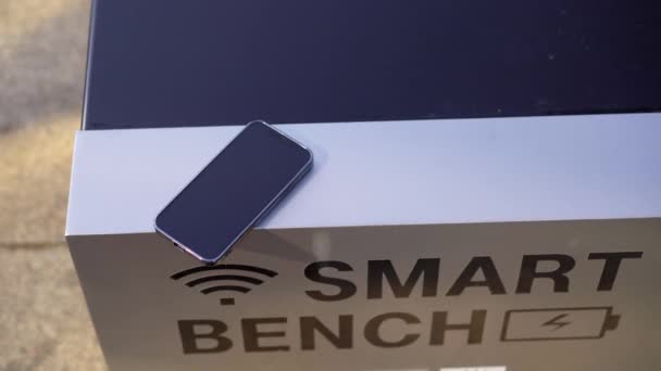 Ένα έξυπνο παγκάκι με ένα smartphone πάνω του. Ένας έξυπνος πάγκος διαθέτει ηλιακούς συλλέκτες στην κορυφή του και μπορεί να φορτίσει δωρεάν συσκευές USB — Αρχείο Βίντεο
