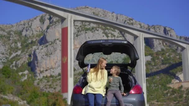 Un uomo e suo figlio sono seduti a mangiare in macchina con il ponte Moracica sullo sfondo. Stanno facendo un viaggio in auto attraverso il Montenegro — Video Stock