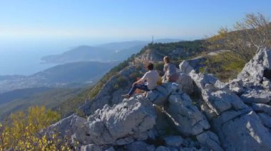 Hava görüntüsü. Turist ailesi, baba ve oğul Karadağ 'daki Kosmach Kalesini ziyaret etti. Uzaktan Budva şehrini izleyen bir tepede dikilirken insansız hava aracı onların etrafında dönüyor.