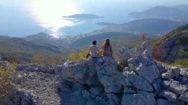 Hava görüntüsü. Turist ailesi, baba ve oğul Karadağ 'daki Kosmach Kalesini ziyaret etti. Uzaktan Budva şehrini izleyen bir tepede dikilirken insansız hava aracı üzerlerinden uçuyor.