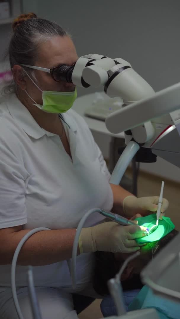 Vídeo vertical. Mulher dentista em um uniforme branco e luvas brancas trata um homem. Ela usa um microscópio para examinar o dente com mais precisão e confiança — Vídeo de Stock