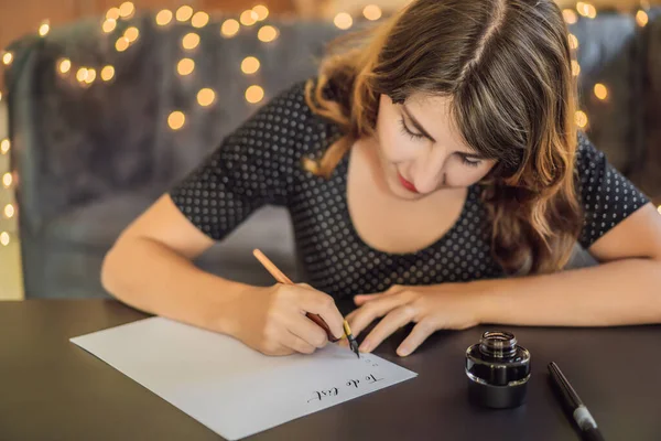 -Hoz csinál oldalra dől. Kalligráfus fiatal nő írja mondat a fehér papíron. Inscribing díszes díszített leveleket. Kalligráfia, grafikai tervezés, betűk, kézírás, létrehozás koncepció — Stock Fotó