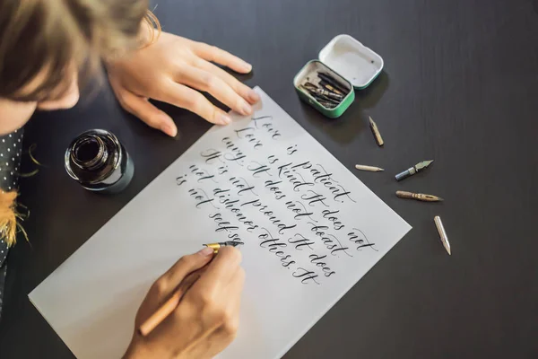 Calígrafo manos escribe frase en papel blanco. Frase bíblica sobre el amor Inscripción de letras ornamentales decoradas. Caligrafía, diseño gráfico, letras, escritura a mano, concepto de creación —  Fotos de Stock