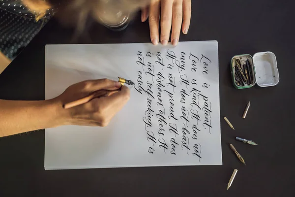 Calígrafo manos escribe frase en papel blanco. Frase bíblica sobre el amor Inscripción de letras ornamentales decoradas. Caligrafía, diseño gráfico, letras, escritura a mano, concepto de creación —  Fotos de Stock