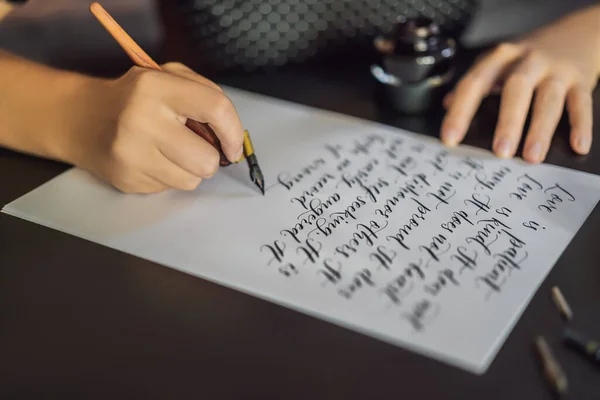 Calígrafo manos escribe frase en papel blanco. Frase bíblica sobre el amor Inscripción de letras ornamentales decoradas. Caligrafía, diseño gráfico, letras, escritura a mano, concepto de creación —  Fotos de Stock