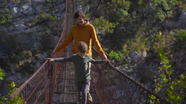 An old metal rope bridge across the Moraca river canyon in Montenegro. A father and son tourists walk along the bridge — Stock Video