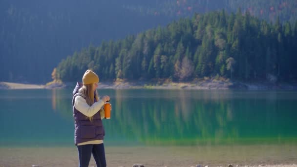 Une jeune femme visite le Crno jezero ou le lac noir près de la ville de Zabljak. Elle boit une boisson chaude dans un thermos au bord du lac. Voyage au nord du Monténégro — Video