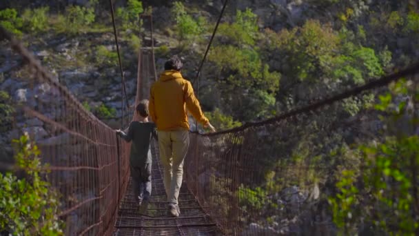 Un viejo puente de cuerda metálica sobre el cañón del río Moraca en Montenegro. Un padre e hijo turistas caminan a lo largo del puente — Vídeo de stock