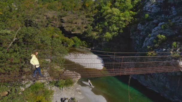 空中ビデオ。モンテネグロのモラカ川渓谷に架かる古い金属製のロープ橋。黄色のジャケットを着た女性が橋を歩いている — ストック動画