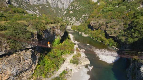 Vídeo aéreo. Un viejo puente de cuerda metálica sobre el cañón del río Moraca en Montenegro. Un hombre con una chaqueta amarilla camina por el puente — Vídeo de stock