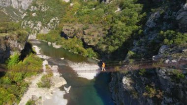Hava videosu. Karadağ 'daki Moraca nehri kanyonu üzerinde eski bir metal ip köprü. Sarı ceketli bir adam köprüde yürüyor.