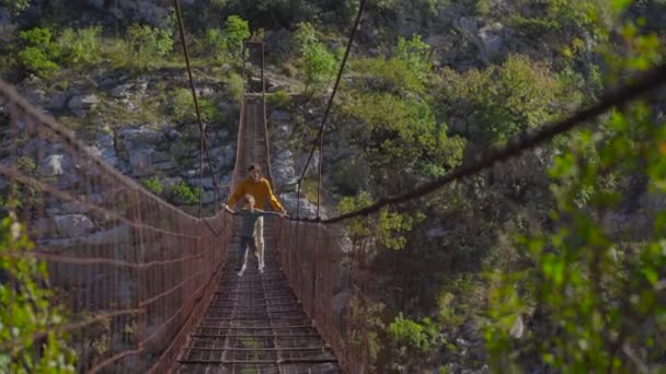 Een oude metalen touwbrug over de Moraca rivierkloof in Montenegro. Een vader en zoon toeristen lopen over de brug — Stockvideo