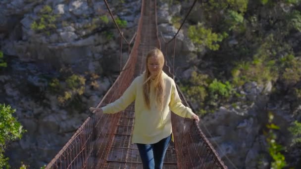 Un viejo puente de cuerda metálica sobre el cañón del río Moraca en Montenegro. Una turista con chaqueta amarilla camina por el puente — Vídeo de stock