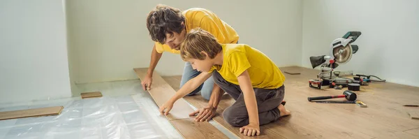 BANNER, LONG FORMAT Ojciec i syn instalują nową drewnianą podłogę laminowaną na ciepłej podłodze filmowej. System ogrzewania podłogowego na podczerwień pod podłogą laminowaną — Zdjęcie stockowe