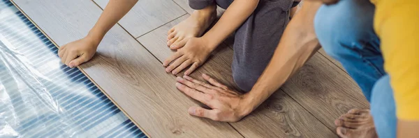 BANNER, LONG FORMAT Vater und Sohn verlegen neuen Holzlaminatboden auf einem warmen Folienboden. Infrarot-Fußbodenheizung unter Laminatboden — Stockfoto