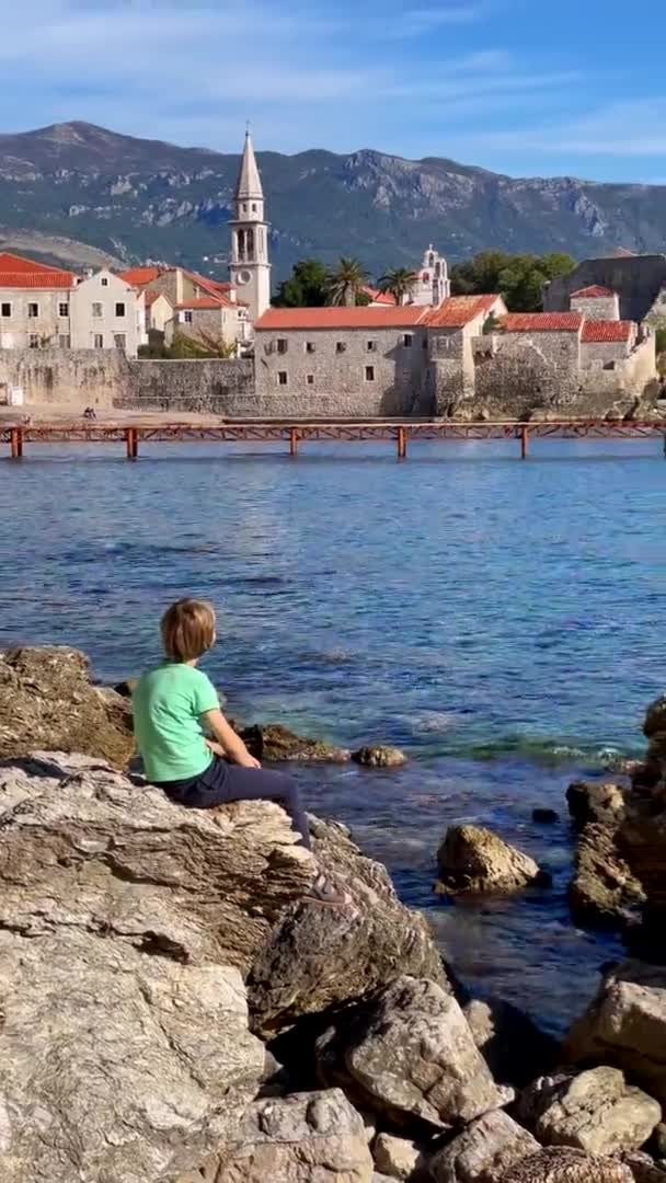 Vídeo vertical. Menino está caminhando ao longo da passarela da praia perto da cidade velha de Budva. Ele se senta em uma rocha observando o forte. Viajar para Montenegro conceito. — Vídeo de Stock