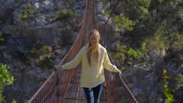 モンテネグロのモラカ川渓谷に架かる古い金属製のロープ橋。黄色のジャケットを着た女性観光客が橋を歩いている — ストック動画