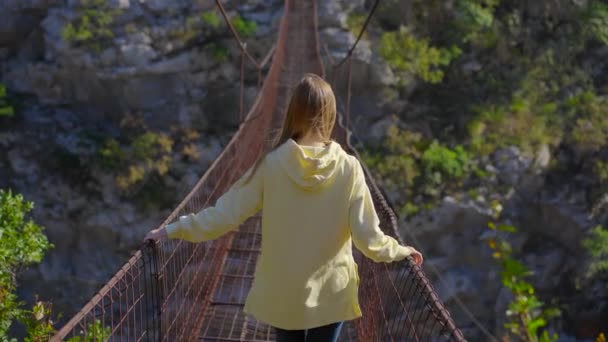 Egy régi fémkötél híd a Moraca folyó kanyonján át Montenegróban. Egy sárga kabátos turista sétál a hídon. — Stock videók
