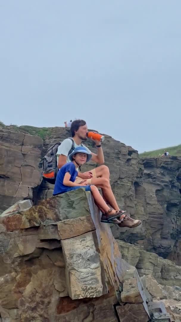 Vertikales Video. Ein junger Mann und sein Sohn sind auf einer Wanderung zwischen Felsen in Meeresnähe. Sie sitzen auf einem Felsbrocken am Meer. Zeitlupenschuss — Stockvideo