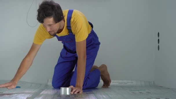 Een professionele meester van vloerverwarmingsinstallatie legt de isolatie voor de vloer uit en lijmt deze vast, waarop hij vervolgens het laminaat zal leggen. — Stockvideo
