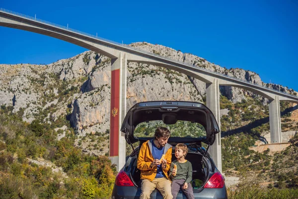 Montenegro. Dad and son tourists are sitting on the trunk of a car. Road trip around Montenegro. Bridge Moracica. Reinforced concrete bridge across the Moraci gorge. The motorway Bar - Bolyare. The
