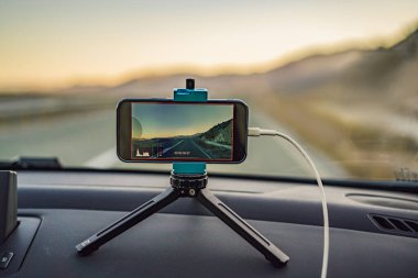 Arabadaki tripodun üzerindeki telefon manzaralı yolun videosunu çeker.