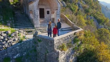 Aerial shot. Family of tourists observes the old town of Kotor standing near the Christian church on a way to the top of the mountain where st. Johns fort is located. Crkva Gospe od Zdravlja, church